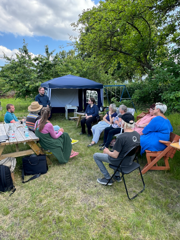 Ein Bild mit Blick in den Garten, wo Menschen im Halbkreis sitzen und David Krüger zuhören.